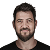 Close-up portrait of a man with dark hair and a beard, wearing a black shirt against a dark background, looking forward.